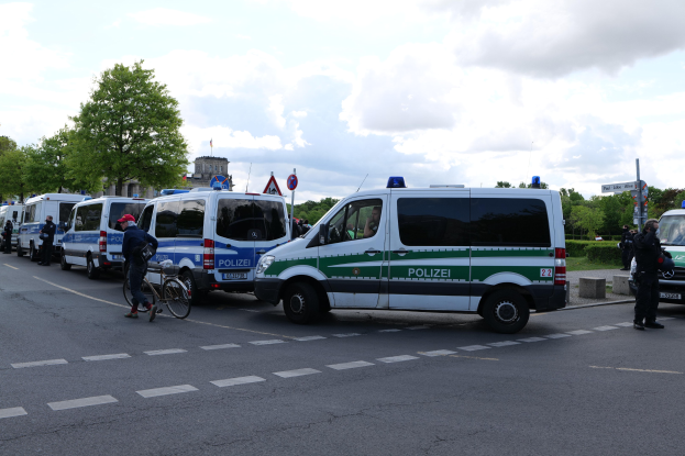 Gruppe von Polizeiwagen entlang einer Straße geparkt mit einem Radfahrer im Vordergrund, Böumen und Gebäuden im Hintergrund unter einem bewölktem Himmel.