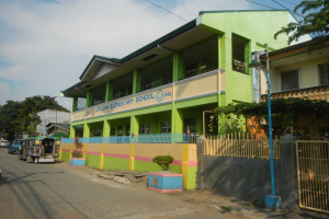Schulgebäude mit grüner und gelber Fassade, Straße mit Fahrzeugen, Topfpflanzen, Zaun, Tor, Straßenlaterne, Bäume und bewölkter Himmel.