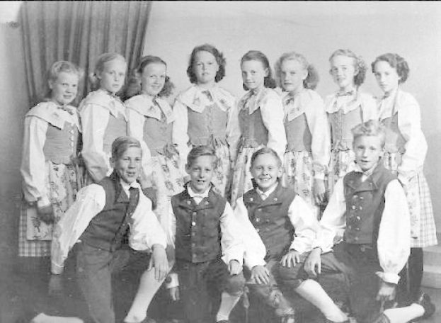 Schwarzes und weißes Foto einer Gruppe von Kindern, die vor einer Wand mit einem Vorhang posieren, beschriftet mit '1930er-Kinder-Schule' unten.