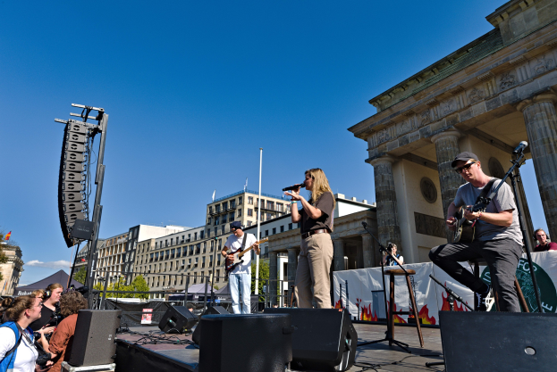 Eine Gruppe von Menschen, die auf einer Bühne Musik spielen, mit dem Brandenburger Tor im Hintergrund, begleitet von Equipment und einer Menge.