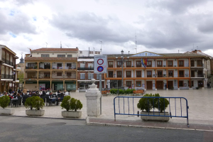 Ein belebter Stadtplatz mit Menschen auf Stühlen sitzend und Stehenden, umgeben von Topfpflanzen, Metallabsperrungen, Straßenmasten mit Flaggen, ein Schild, Gebäude und ein bewölkter Himmel.