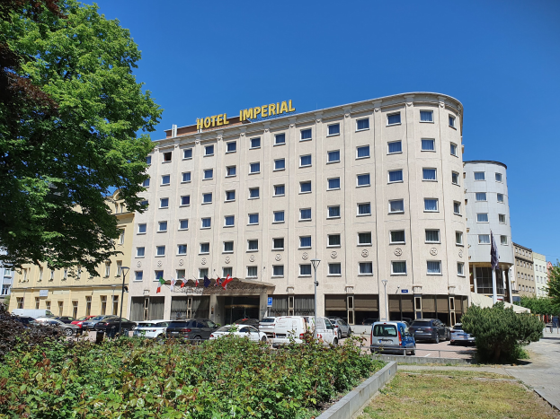 Das Hotel Imperial in Berlin, Deutschland, ein großes Gebäude mit Fenstern, umgeben von Grün, parkenden Autos, Straßenlaternen mit Flaggen, unter einem bewölkten Himmel.