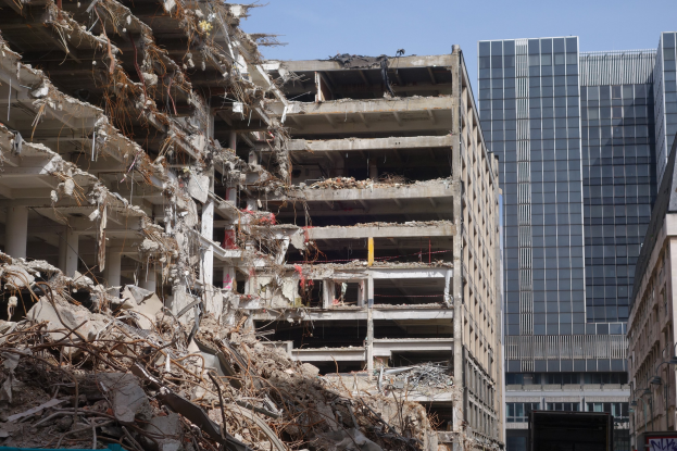 Ein großes Gebäude wird in einer von anderen Gebäuden, Fahrzeugen, Straßen-Elementen, Bäumen und einem klaren Himmel umgebenen Stadt abgerissen.