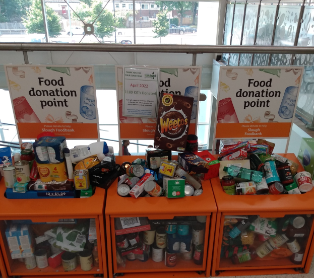 Orange Schrank mit Regalen voller Flaschen, Dosen, Schachteln und anderen Gegenständen, mit Informationsschildern; Glasfenster im Hintergrund zeigen Fahrzeuge, Bäume, Gebäude und Himmel.