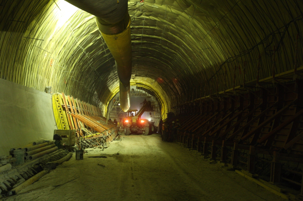 Baustelle mit einem großen Tunnel, Fahrzeugen, verstreuten hölzernen Gegenständen, Rohren, einer Wand auf der linken Seite und beleuchtetem Hintergrund.
