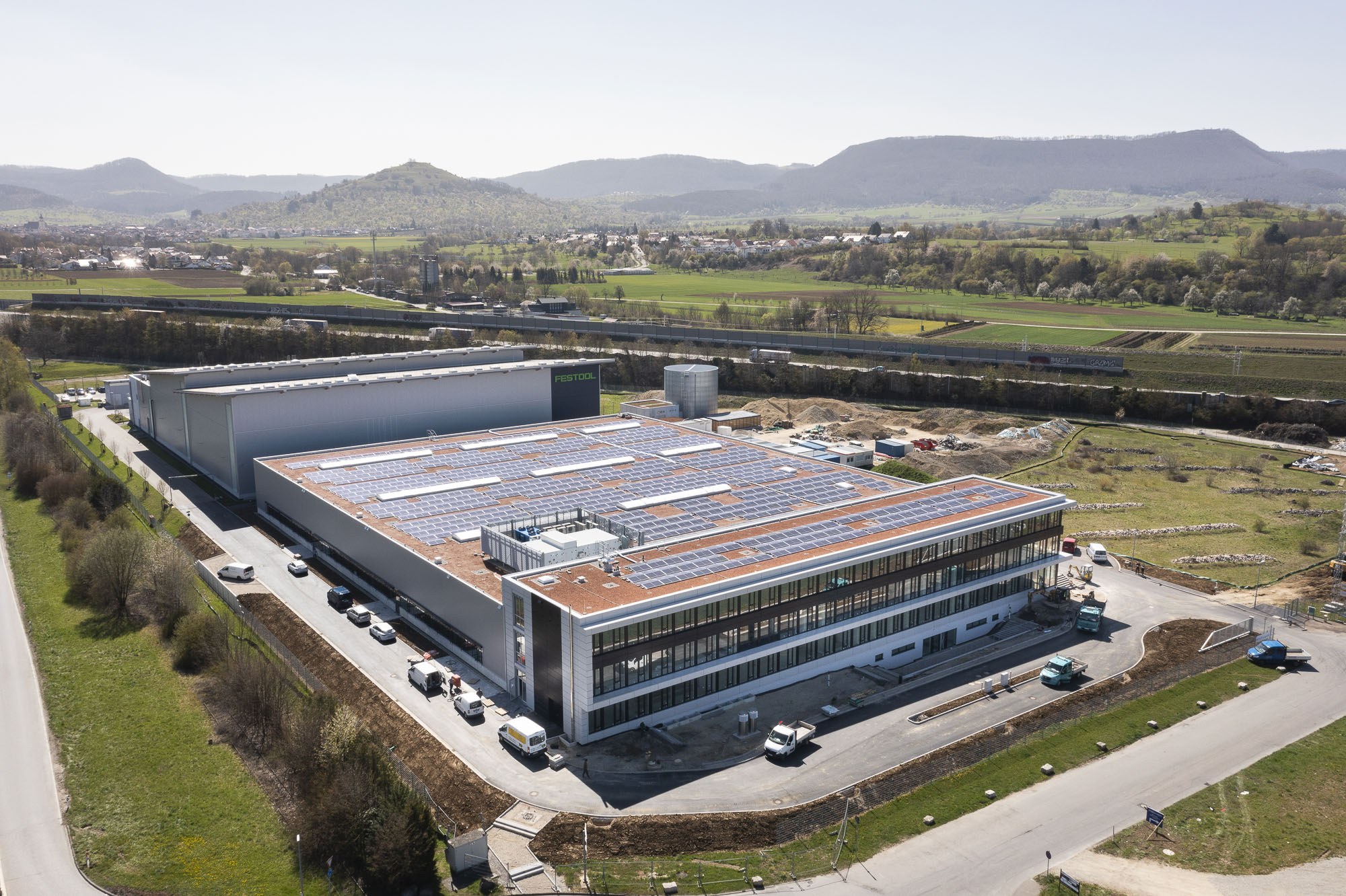 Luftaufnahme eines große Fabriks mit Solarmodulen auf dem Dach, umgeben von Fahrzeugen, Bäumen, Gras und Strommasten, vor Hühgeln und einem klaren blauen Himmel.