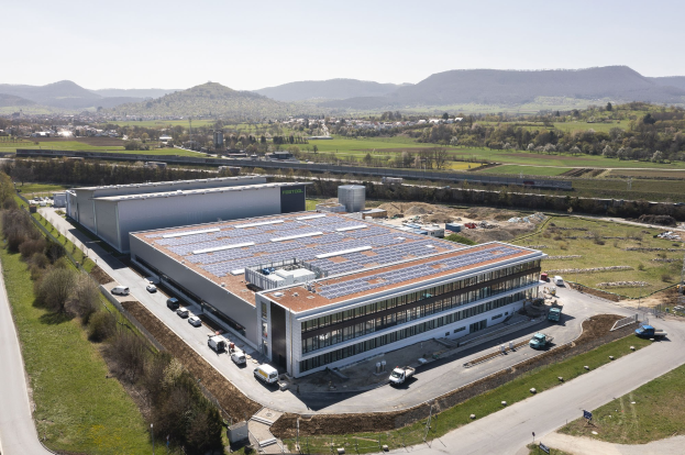Luftaufnahme eines große Fabriks mit Solarmodulen auf dem Dach, umgeben von Fahrzeugen, Bäumen, Gras und Strommasten, vor Hühgeln und einem klaren blauen Himmel.