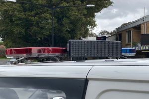 Ein Polizeiwagen mit eingeschaltetem Blaulicht steht an der Straße geparkt, umgeben von Gebäuden, Bäumen, Polen und Schildern, mit einem bewölkten Himmel im Hintergrund, wahrscheinlich nach einem tödlichen Schulschießen in Brisbane.