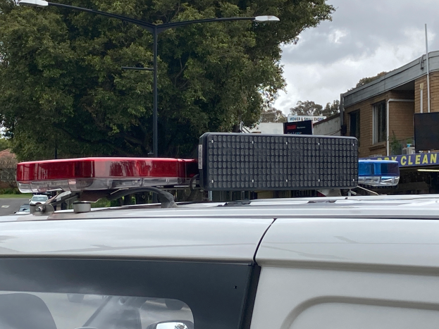 Ein Polizeiwagen mit eingeschaltetem Blaulicht steht an der Straße geparkt, umgeben von Gebäuden, Bäumen, Polen und Schildern, mit einem bewölkten Himmel im Hintergrund, wahrscheinlich nach einem tödlichen Schulschießen in Brisbane.
