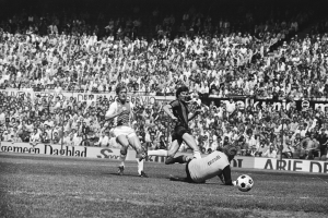 Schwarzes und weißes Foto von Männern, die auf einem Feld Füßball spielen, mit Zuschauern im Hintergrund und einer Bundesliga 1978-79 Ajax 1-1 Hannover 96 Fahne.