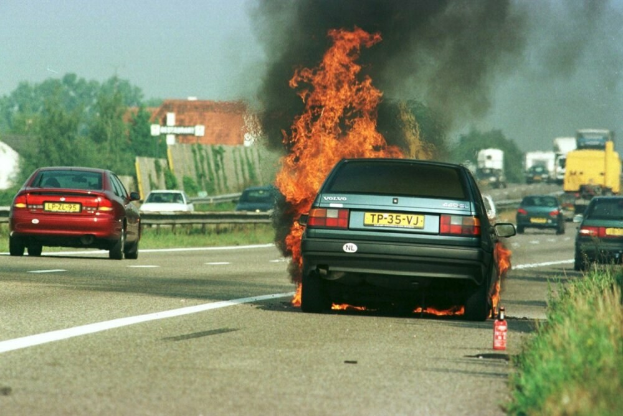 Ein Auto, das in Flammen steht, am Straßenrand mit anderen Fahrzeugen in der Nähe, Bäume und Gebäude im Hintergrund unter einem klaren blauen Himmel und Gras mit einem Feuerlöscher auf der rechten Seite.