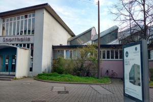 Ein großes Gebäude mit einem Schild davor, umgeben von Bäumen, Pflanzen, Gras, Fahrrädern und einem Pfahl, unter einem bewölkten Himmel, identifiziert als Sportballe, zur Eröffnung 2020 vorgesehen.