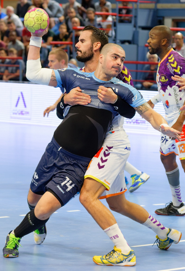 Eine Gruppe von Männern, die Handball in einem Stadion spielen, wobei einer der Spieler den Ball hält.