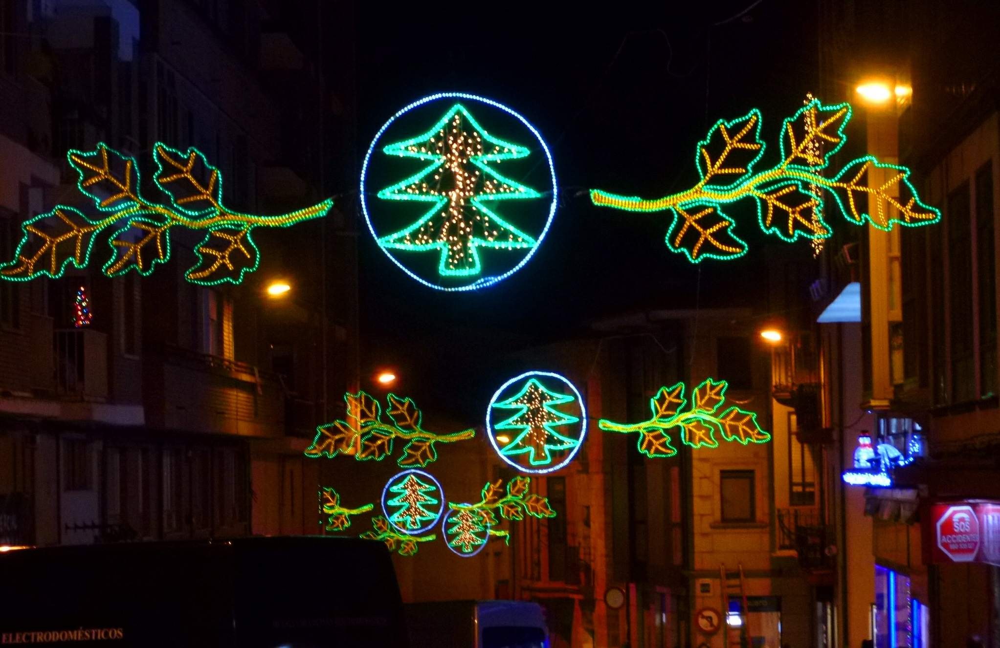 Eine städtische Straße, die mit festlichen Weihnachtslichtern geschmückt ist, die Blätter und andere Verzierungen zeigen, mit Fahrzeugen, die an Gebäuden auf beiden Seiten vorbeifahren.