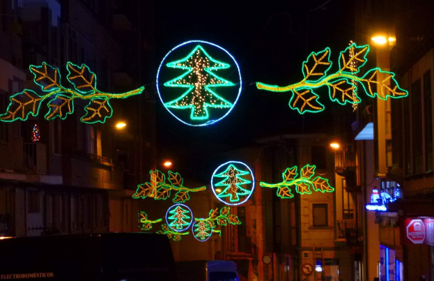 Eine städtische Straße, die mit festlichen Weihnachtslichtern geschmückt ist, die Blätter und andere Verzierungen zeigen, mit Fahrzeugen, die an Gebäuden auf beiden Seiten vorbeifahren.