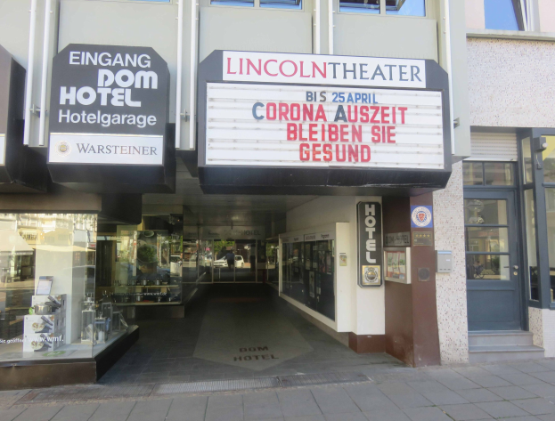 Außenansicht des Lincoln Theaters in Berlin, Deutschland, mit Glasfenstern, Türen und einer Texttafel, wobei eine belebte Stadtlandschaft im Inneren zu sehen ist.
