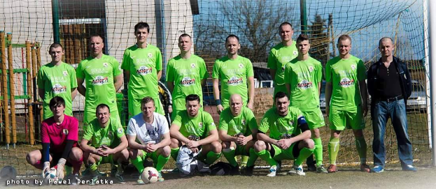 Eine Gruppe von Männern in grünen T-Shirts steht und hockt auf einem Fußballfeld mit einem Ball davor, ein Torpfosten rechts daneben und ein Hintergrund aus Bäumen, Gebäuden, Pfählen, Fahrzeugen und dem Himmel.