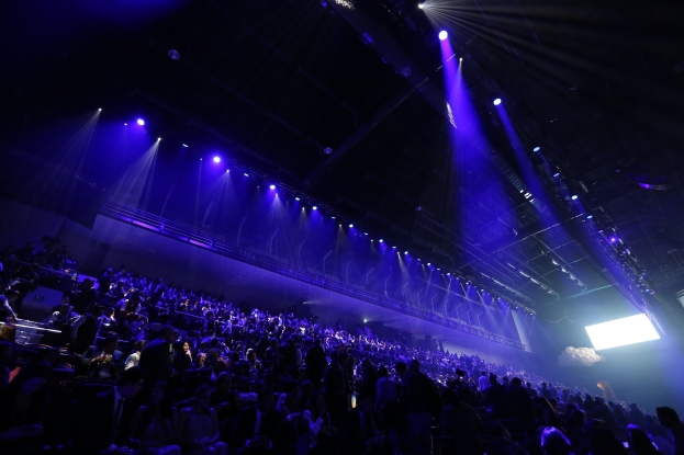 Große Menschenmenge in einem abgedunkelten Konzertsaal, der einer Bühne mit Deckenlampen und einem Bildschirm auf der rechten Seite gegenüberliegt.