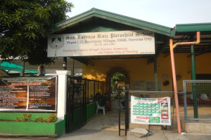 Der Eingang der San Lorenzo Ruiz Pfarrschule hat ein Schild mit Text, einen Metallzaun, Menschen, die auf Stühlen unter einem Dach sitzen, Pfosten, ein Hinweisschild, Pflanzen, einen Baum und einen bewölkten Himmel.