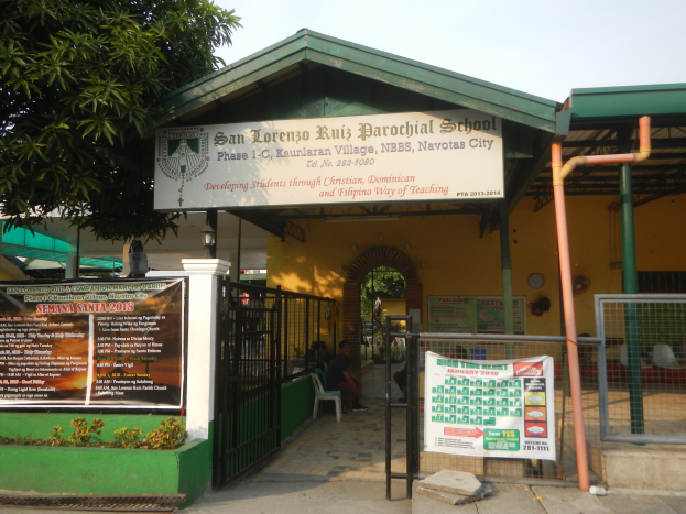 Der Eingang der San Lorenzo Ruiz Pfarrschule hat ein Schild mit Text, einen Metallzaun, Menschen, die auf Stühlen unter einem Dach sitzen, Pfosten, ein Hinweisschild, Pflanzen, einen Baum und einen bewölkten Himmel.