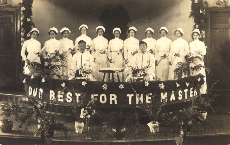 Gruppe von Frauen in weißen Kleidern und Kappen auf einer Bühne mit einem zentralen Banner, auf dem "Unser Bestes für den Meister" steht, umgeben von Blumentöpfen und vor Türen und einer Wand.