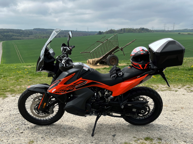 Eine KTM 1290 Super Adventure R Motorrad auf einem Schotterweg mit Helm und Gepäckstück daneben, umgeben von grünem Gras, hohen Bäumen und Strommästen unter einem bewölktem Himmel.