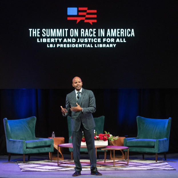 Ein Mann in einem Blazer, Krawatte und Hemd steht auf einer Bühne und spricht bei einem Event mit dem Titel "The Summit on Race in America: Liberty and Justice for All" in der LBJ Presidential Library, mit zwei Stühlen, einem Tisch mit einem Blumenstrauß und einer Flasche darauf und einem Bildschirm dahinter.