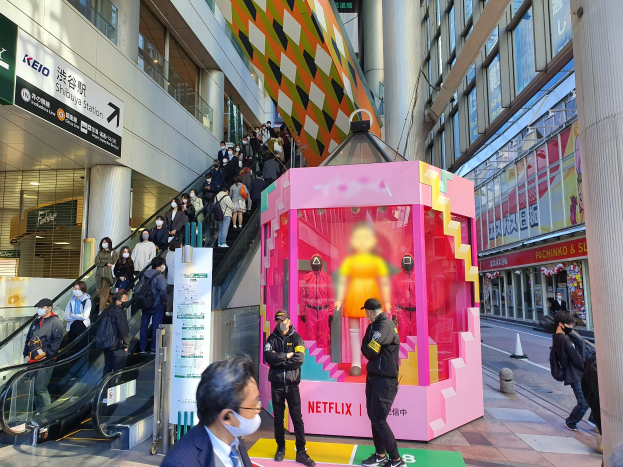 Eine Gruppe von Menschen steht vor einem modernen Gebäude mit Glaswänden und Säulen, einige tragen Masken, und eine Rolltreppe ist zu sehen. Eine pinkfarbene Box in der Mitte zeigt Schaufensterpuppen und trägt die Aufschrift "Netflix's new Netflix booth in Tokyo."