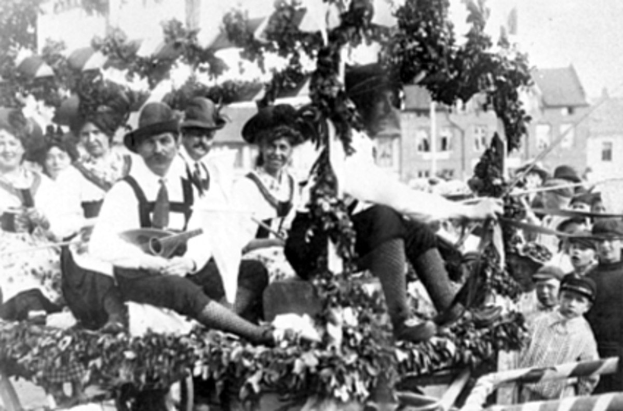 Schwarzes und weißes Bild eines Paradewagens mit Menschen in traditioneller bayrischer Tracht, einige spielen Instrumente, mit Blattdekoration, vor einem Hintergrund aus Gebäuden und einem klaren Himmel.