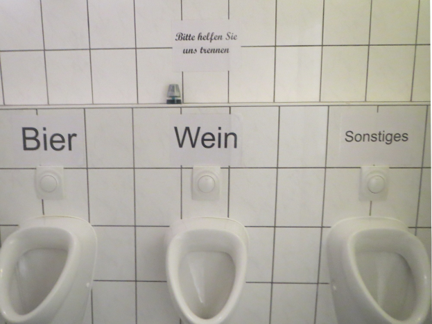Drei weiße Toiletten an einer Wand montiert mit Überschriften über jeder, die "Bier Wein" und "Sonstiges" lesen.