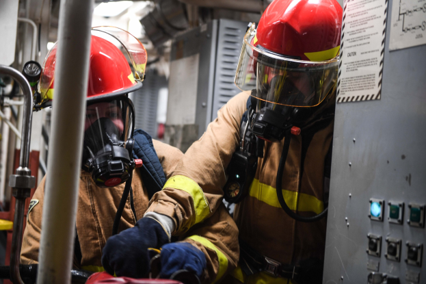 Zwei Feuerwehrleute in Schutzausrüstung, Helmen und Gasmasken arbeiten an einem Feuerwehrauto mit einer Tafel und Text auf der rechten Seite und Metallstangen im Hintergrund.