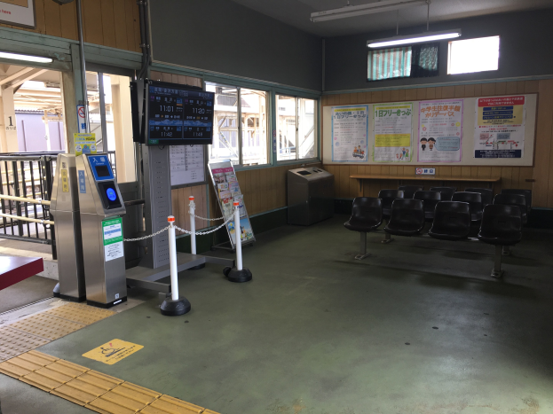 Ein belebter Bahnhofseingang mit Sitzgelegenheiten, einer Informations counter, Ticketautomaten, Kettenabsperrungen, Informationsanzeigen, Wandplakate, Deckenbeleuchtung und Geländern.