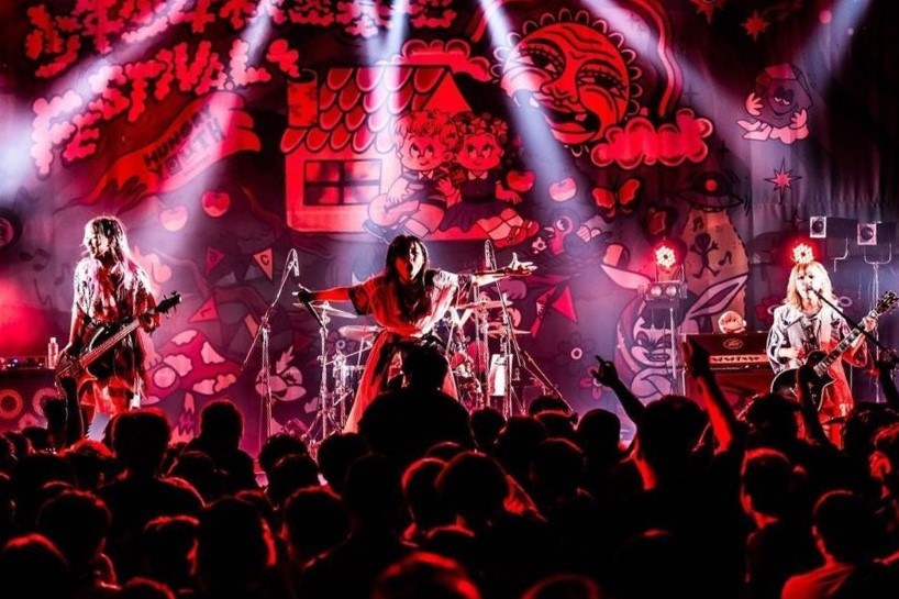 Ein Live-Auftritt der japanischen Rockband Korn auf der Bühne, mit Bandmitgliedern, die Gitarren spielen und in Mikrofone singen, einem beleuchteten Gemälde an der Wand dahinter und einem sichtbaren Publikum unten.
