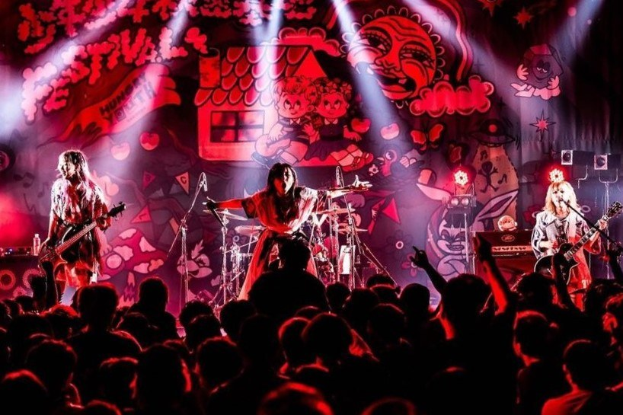 Ein Live-Auftritt der japanischen Rockband Korn auf der Bühne, mit Bandmitgliedern, die Gitarren spielen und in Mikrofone singen, einem beleuchteten Gemälde an der Wand dahinter und einem sichtbaren Publikum unten.
