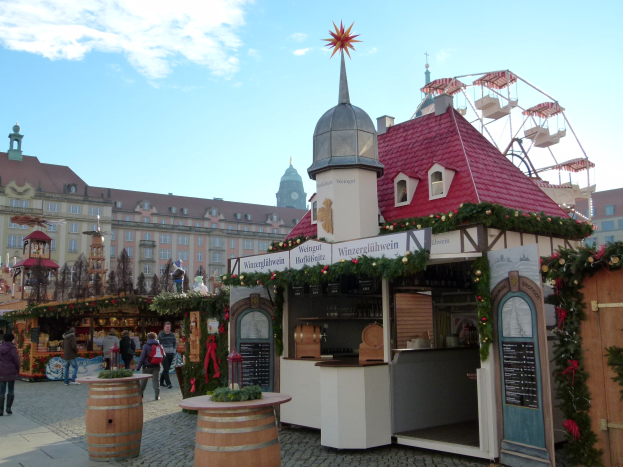 Ein geschäftiger Weihnachtsmarkt in Nürnberg, Deutschland, mit Menschen um dekorierte Stände, festlicher Beleuchtung und Schmuck, Gebäuden im Hintergrund, einem Riesenrad und einer Tafel auf der rechten Seite.