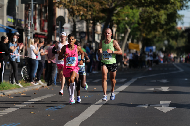 Gruppe von Menschen, die bei einem Marathon auf einer Stadtstraße laufen, mit Zuschauern, unscharfen Bäumen, Gebäuden und einem Fahrrad im Hintergrund.
