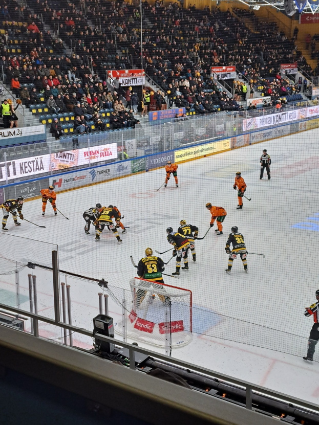 Gruppe von Menschen, die Hockey auf einem Eisring spielen, umgeben von einem Zaun, mit einem Netz in der Mitte, Zuschauern im Hintergrund und beleuchteten Stadionlampen oben.