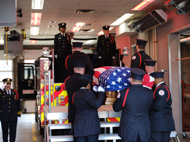 Eine Gruppe uniformierter Männer trägt einen Sarg mit einer amerikanischen Flagge bei einer Beerdigungszeremonie, umgeben von Menschen mit Mützen und einem Löschfahrzeug sowie Deckenleuchten im Hintergrund.