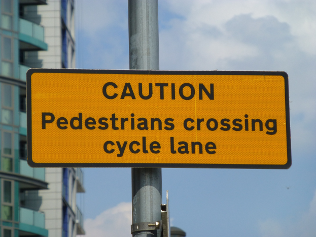 Gelbes Warnschild an einem Pfahl vor einem Gebäude, auf dem steht "Fußgänger überqueren Fahrradspur".