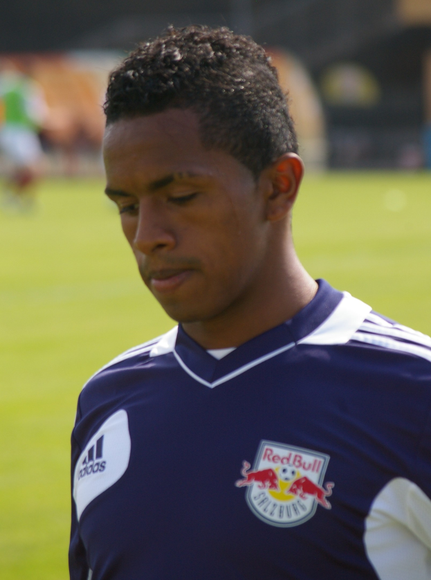 Junger Mann in einem blauen T-Shirt auf einem Fußballfeld stehend, der wie ein Flügelstürmer mit kurzen, dunklen Haaren und einem entschlossenen Gesichtsausdruck aussieht.