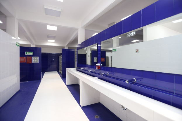Öffentliche Toilette mit blauen Kacheln, weißen Tresen, Waschbecken mit Wasserhähnen, einer Plattform mit einer Flasche und anderen Gegenständen, Texttafeln an den Wänden, Deckenleuchten und Stützpfeilern.