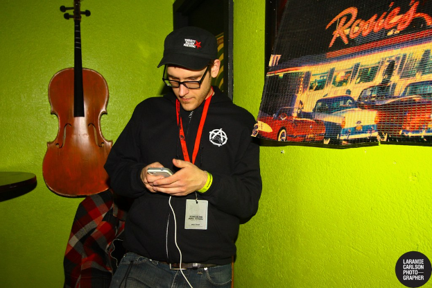 Eine Person mit einer Mütze und einem Ausweis hält ein Mobiltelefon in der Hand, mit einer Gitarre links und einem Plakat rechts.