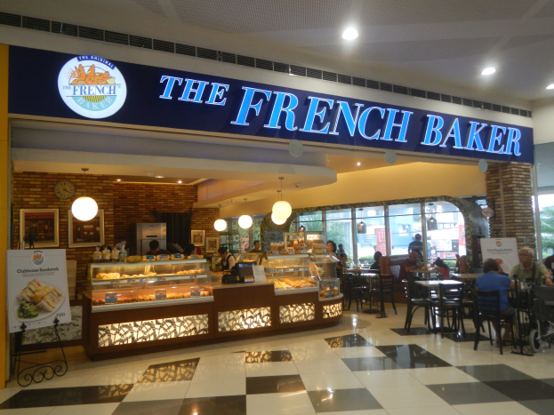 Ein Bäckerei namens 'The French Baker' im Mall of America mit sitzenden und stehenden Menschen, Tischen mit Gegenständen, einer Auslage, Schildern, einer Uhr, gerahmten Fotos an der Wand, Deckenleuchten und Lüftern.