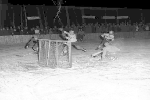Schwarz-weißes Bild eines Hockey-Spiels auf einem Eisstadion, mit Spielern in der Nähe eines Tores und Zuschauern im Hintergrund.