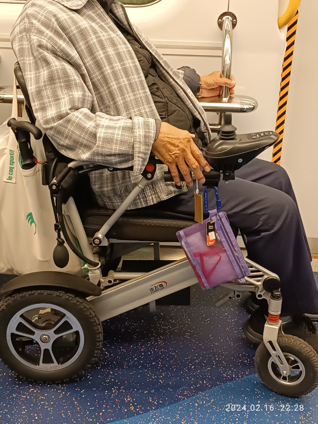 Ein älterer Mann im Rollstuhl in einem Zug, mit einer Tasche neben ihm und einem Spiegel an der Wand dahinter.