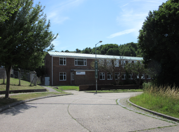 Ein Schulgebäude mit Fenstern, ein Straßenschild, ein Schild, eine Gruppe von Bäumen, Gras, ein Zaun und ein bewölkter Himmel, das in der Mitte eines Parks mit saftigem Gras liegt.