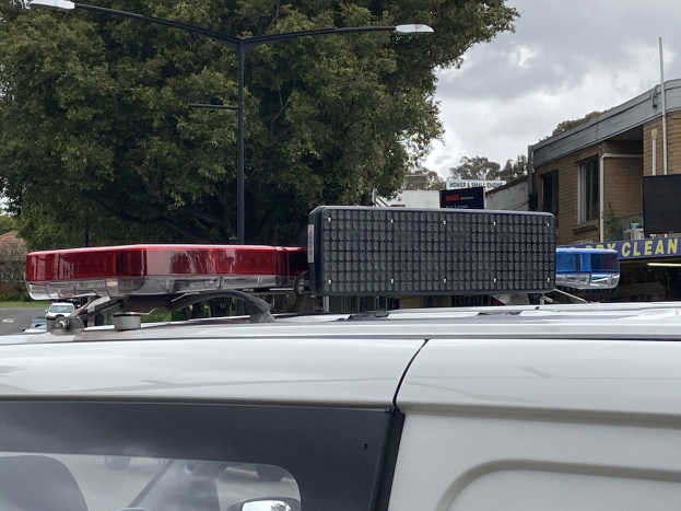 Ein Polizeiwagen mit blinkenden Lichtern steht an einer Straße neben Gebäuden und Bäumen, mit Schildern und Strommasten sichtbar, unter einem bewölkten Himmel.