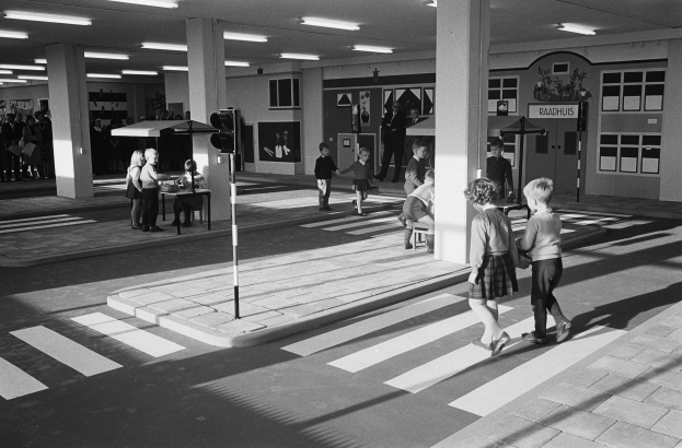 Schwarzes und weißes Foto von Kindern, die eine Straße an einer Ampel überqueren, mit Fußgängern und einer Verkehrsampel im Vordergrund und Säulen mit Deckenleuchten im Hintergrund.