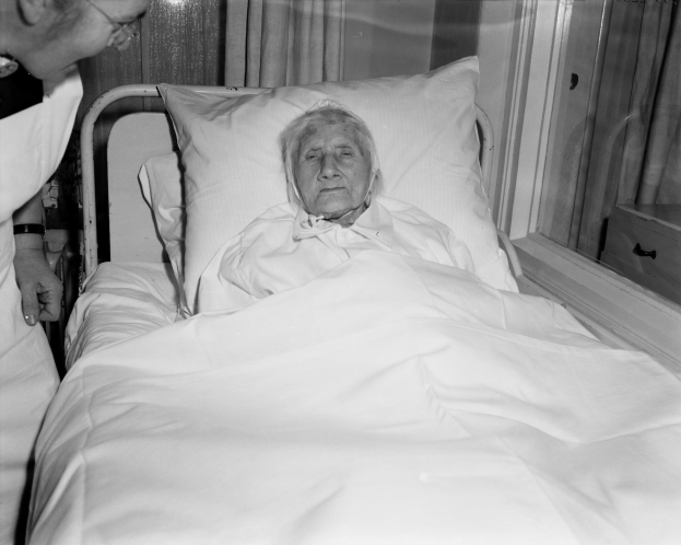 Ein älteres Weib liegt in einem Krankenhausbett mit einer weißen Decke und Kissen, während eine Krankenschwester neben ihr steht, mit einem Fenster und Vorhängen im Hintergrund.