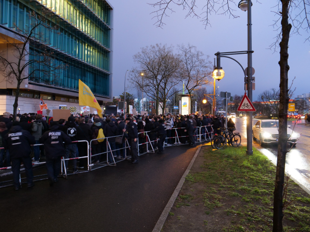 Eine große Gruppe von Menschen mit Schildern steht vor einem Gebäude mit Barrikaden, Fahrrädern, Laternen, Schildern, Bäumen und Gras, mit dem Himmel im Hintergrund, was auf eine Demonstration in Berlin hinweist.